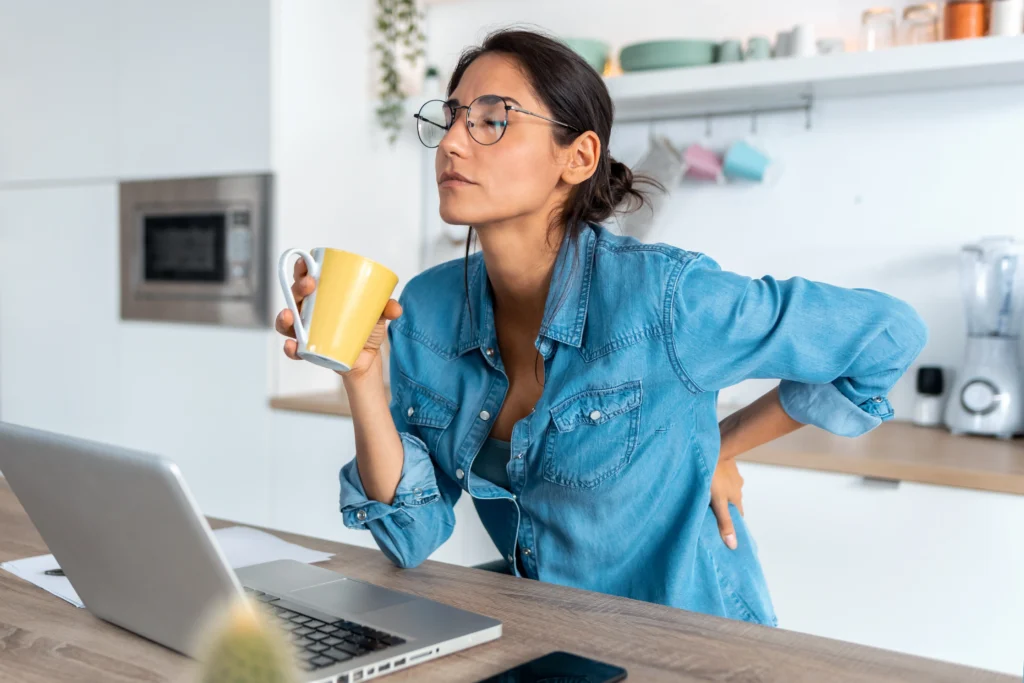 Women holding her lower back while at her computer and drinking coffee. She looks in lower back pain.