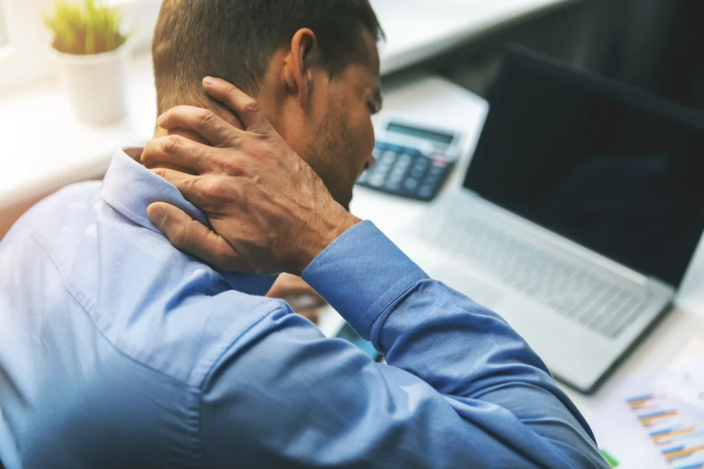 View of a Man at work desk from behind/ right-side. He is grabbing his neck as if it is in pain while he is working. Breaking the Pain Cycle with Chiropractic Care.