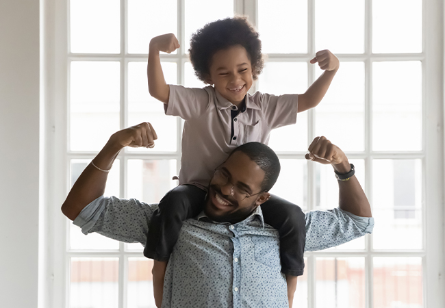father smiling with son on his shoulders both flexing biceps.