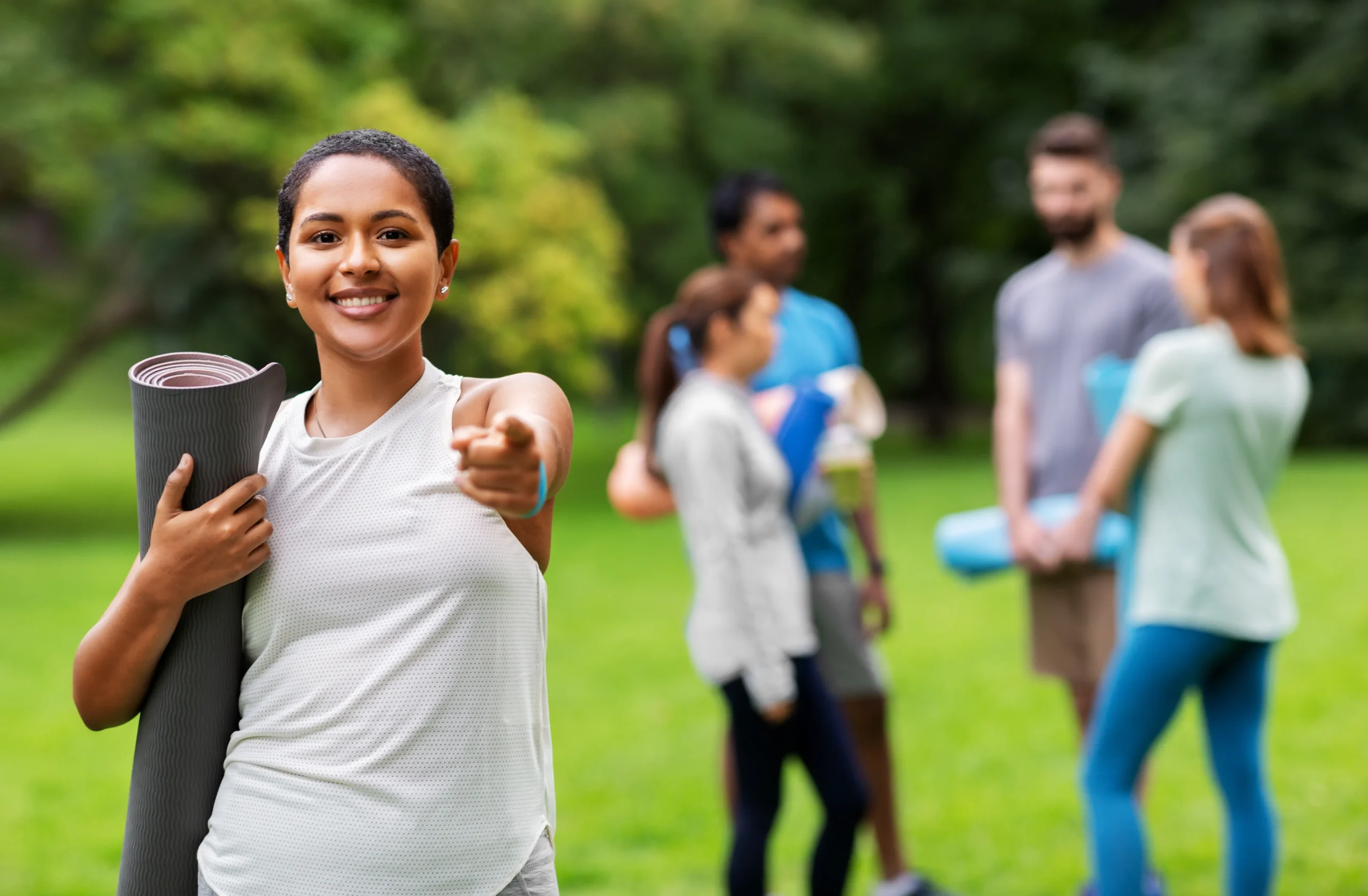 A woman stands in a park, with a group of others behind her. She's holding a yoga mat, ready for a session focused on spinal alignment and wellness.